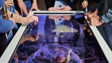 Several people are standing at a touchscreen table. On the left, someone is showing something on the screen.