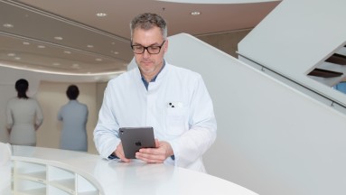 A doctor in white scrubs is holding an Apple tablet, with two healthcare professionals standing in the background in a clinic 