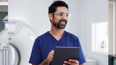 A man is in the radiology examination room, wearing a blue medical gown and glasses, and holding a tablet in his hands.